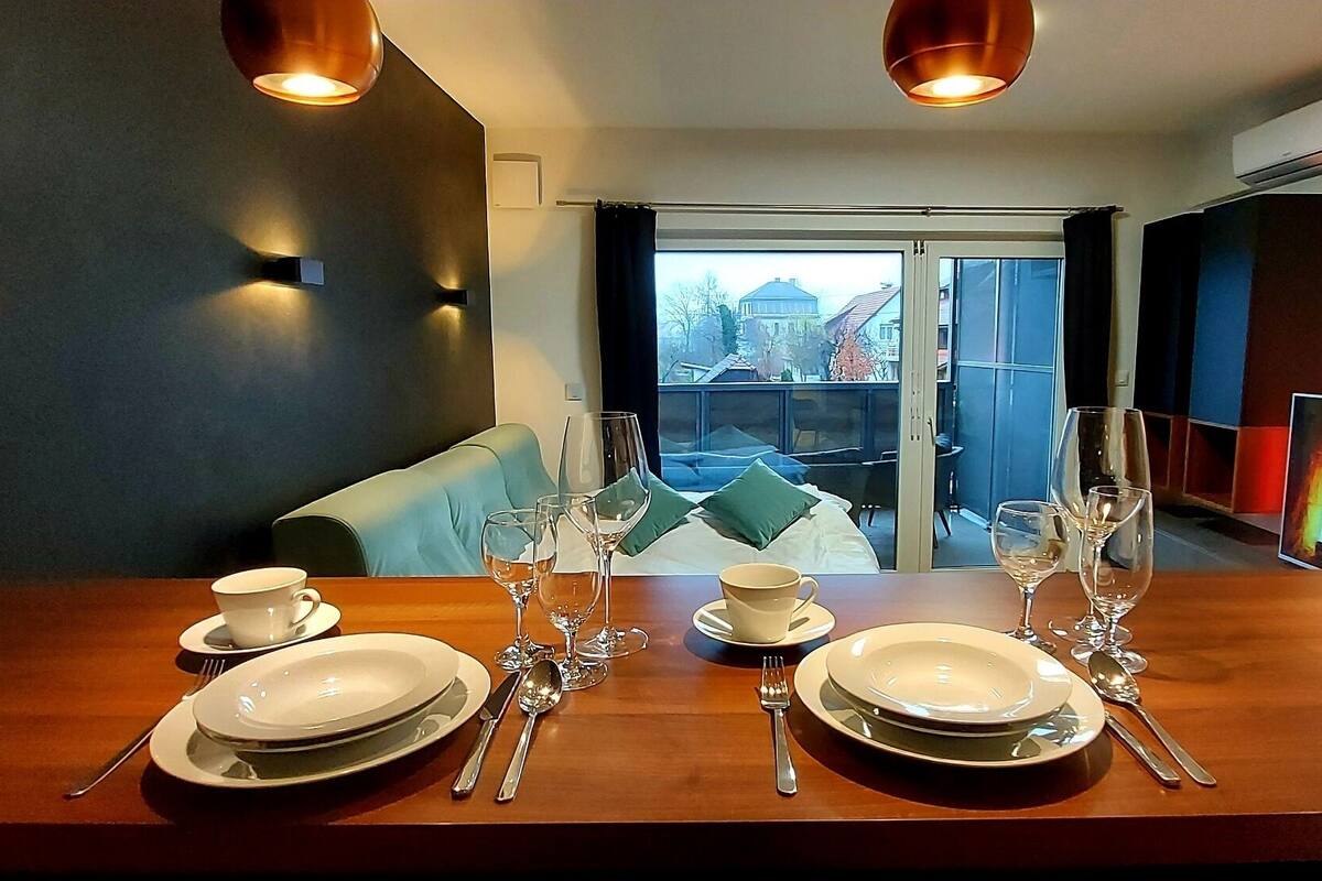 A dining area with a wooden table set for four, including plates, cutlery, and wine glasses. There is a sofa with cushions in the background, and a television displaying a fire scene is mounted on the wall.