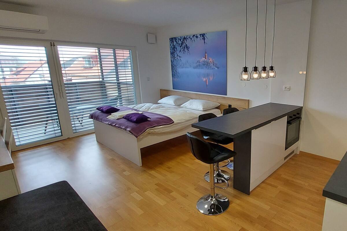 A modern bedroom with a bed, a small desk, and a chair. There is a window with blinds, a painting on the wall, and a kitchen area with a counter and appliances.