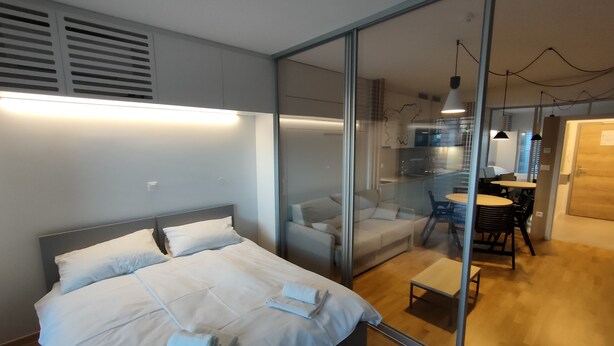 A modern hotel room with a bed, a glass partition, a dining area, and a kitchen visible through the glass. - REZIDENCA PIPANOVA (Ljubljana)