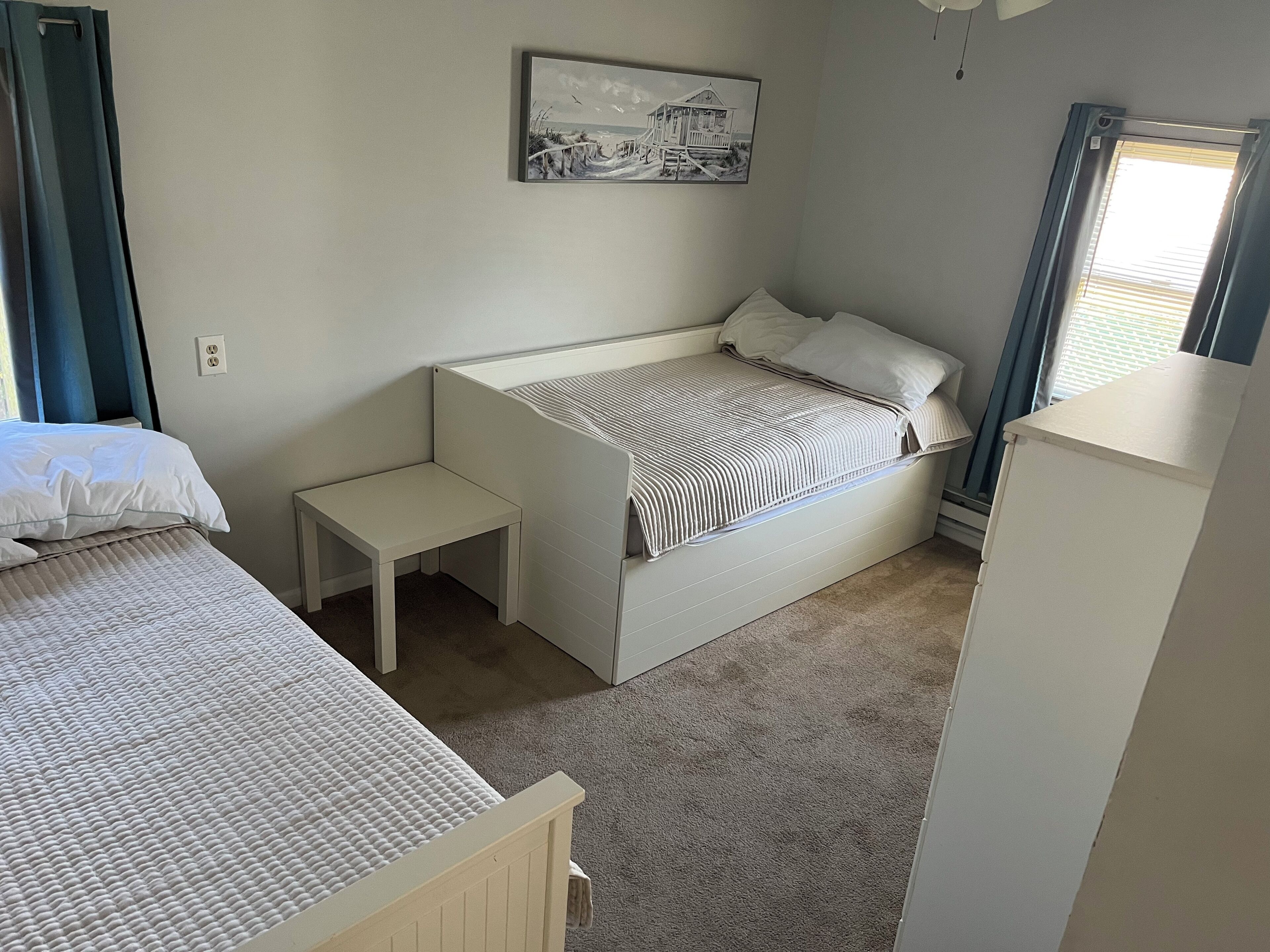A bedroom with two beds, a small bedside table, a headboard, a window with blinds, and a wall-mounted picture.
