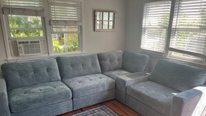 A grey sectional sofa with a patterned rug, large windows with blinds, and an air conditioning unit. - Beach Haven Long Beach Island (Beach Haven)