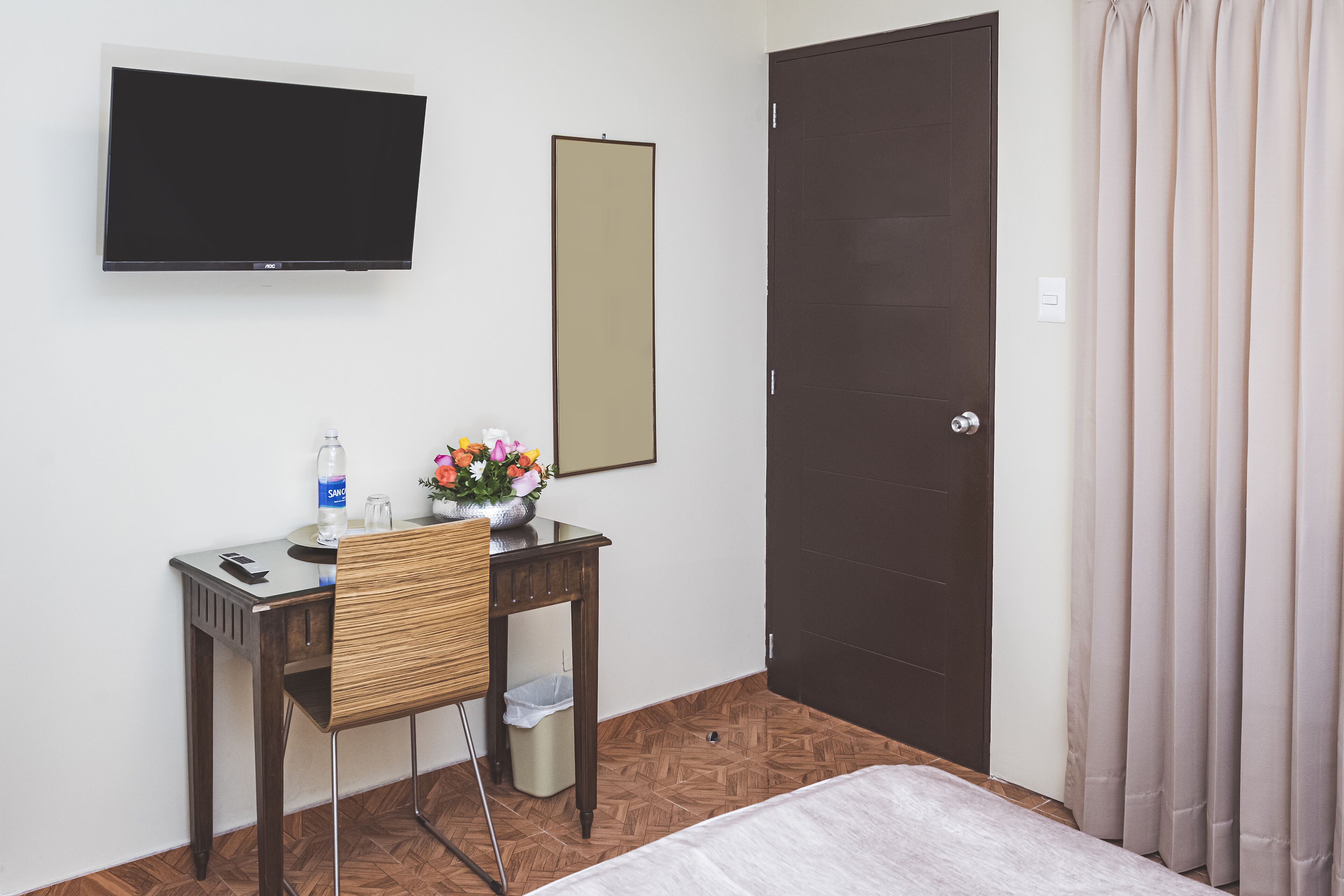 A hotel room with a bed, a desk with a chair, a TV mounted on the wall, a mirror, and a door with curtains.
