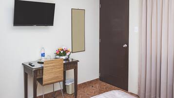 A hotel room with a bed, a desk with a chair, a TV mounted on the wall, a mirror, and a door with curtains.