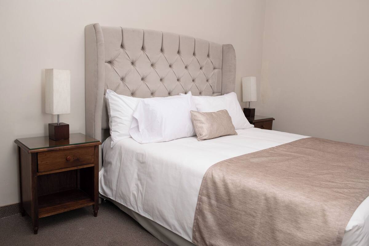 A neatly made bed with a tufted headboard, white bedding, and a brown throw blanket. There are two bedside tables with lamps and a small drawer.