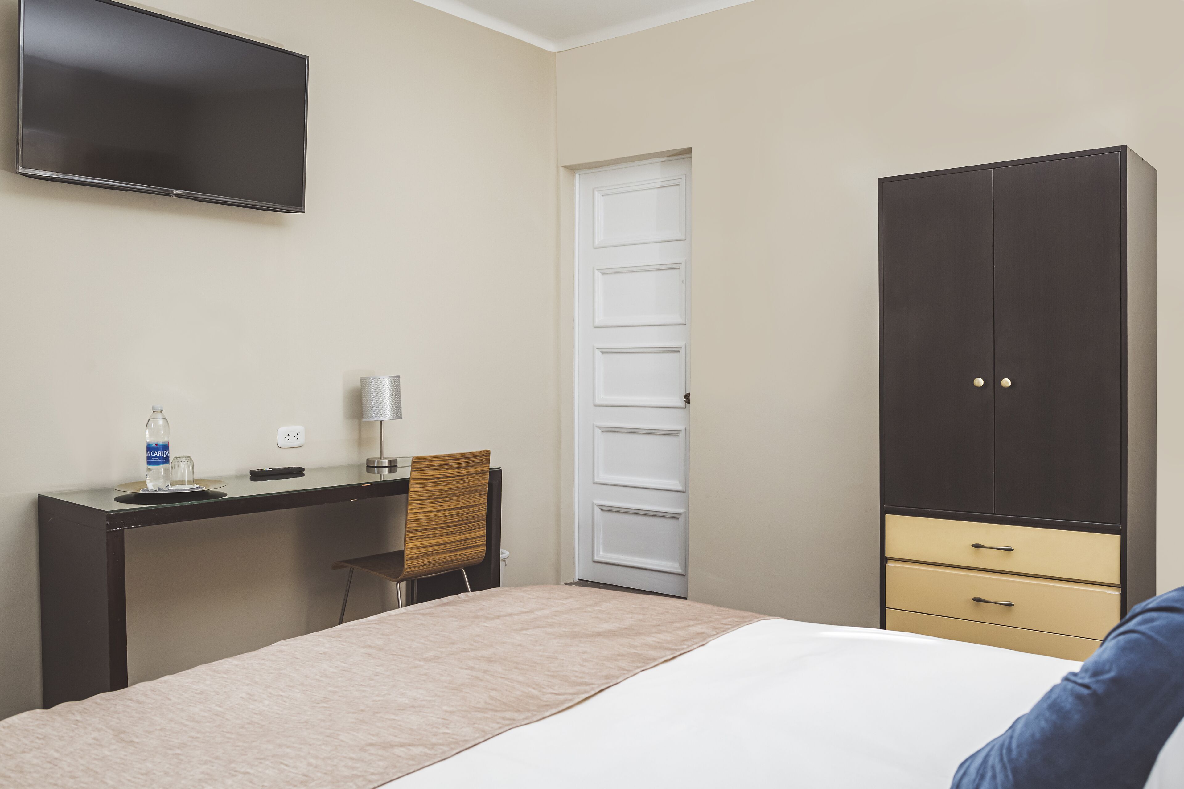 A hotel room with a bed, a desk with a chair, a wardrobe, and a television mounted on the wall.