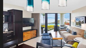 Een moderne woonkamer met een bank, salontafel en televisie. Er is een keuken met kastjes en een waterkoker. De kamer heeft een groot raam met uitzicht op zee.
