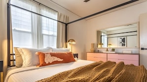 Una habitación de hotel con una cama, un ventanal grande, una mesita de noche con lámparas y un aparador.