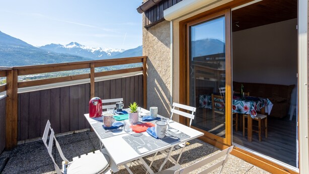 En balkon med et bord dækket til to, en rød elkedel og en potteplante.