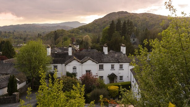 A white building with multiple chimneys, surrounded by lush greenery and hills in the background.