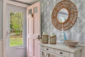Une salle de bain avec un meuble-lavabo blanc, une porte rose, un miroir avec un cadre tressé et un papier peint à motifs.