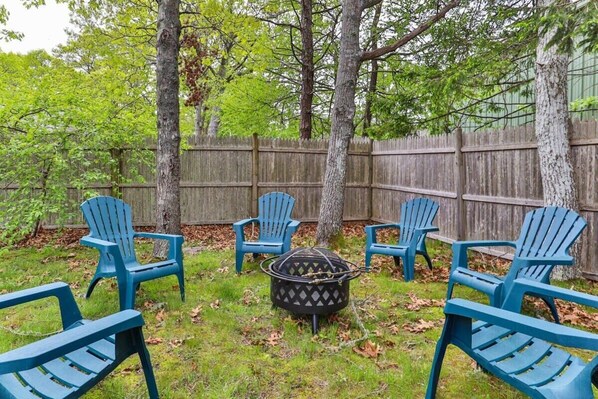 Patio trasero con un brasero rodeado de sillas Adirondack azules, una valla de madera y árboles.