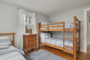Una habitación con una cama nido, un aparador de madera, una ventana con persianas y una alfombra gris.