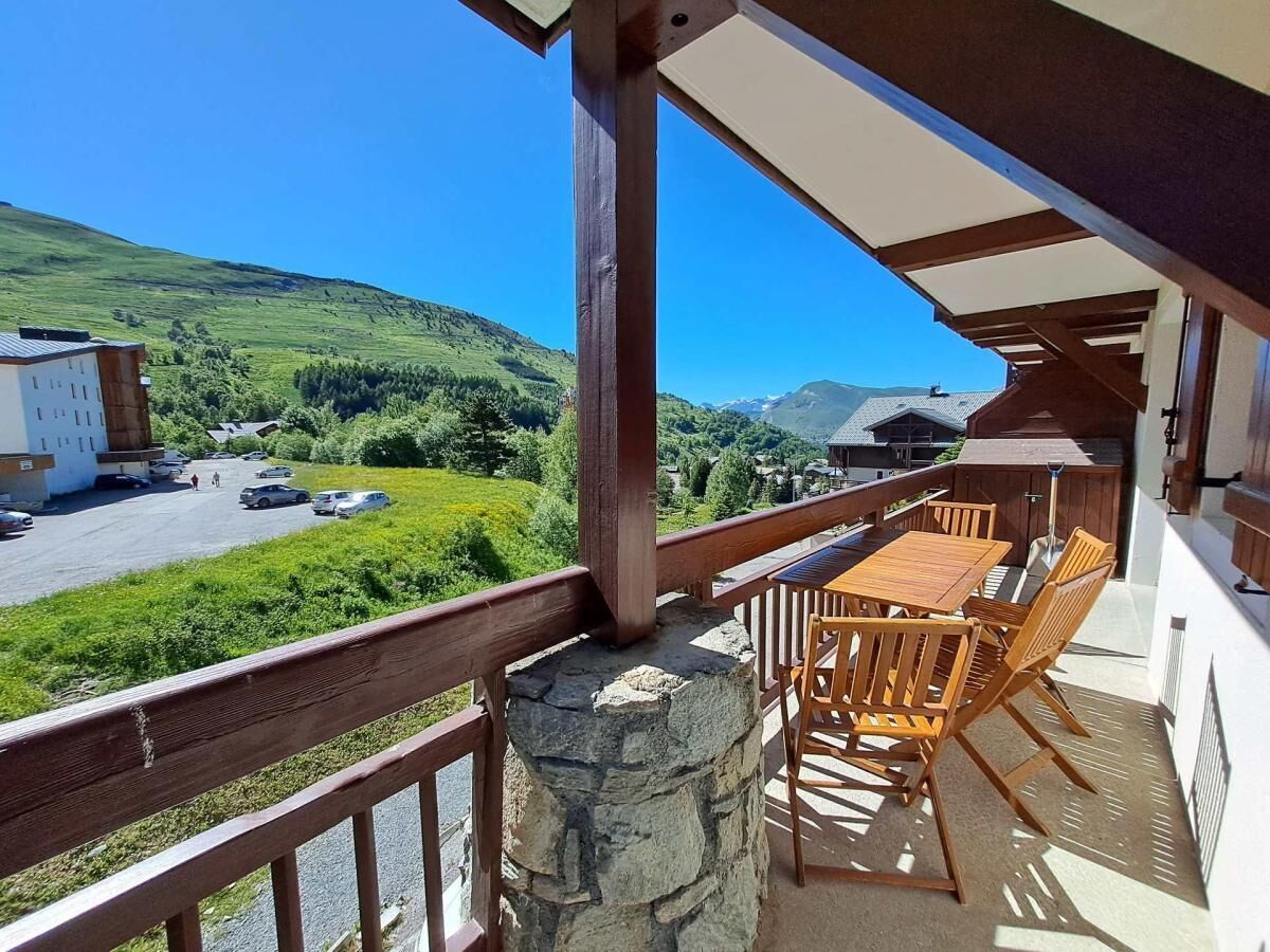 A balcony with a stone pillar, wooden table and chairs, and a view of a parking area and green landscape.