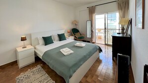 A bedroom with a bed, nightstands, a chair, a desk, and a view of the outside.