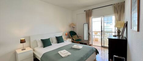 A bedroom with a bed, nightstands, a chair, a desk, and a view of the outside.