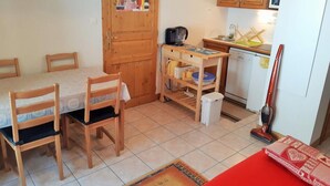 Cocina compacta con armarios de madera, una mesa de comedor con sillas, microondas, aspiradora y un refrigerador pequeño.