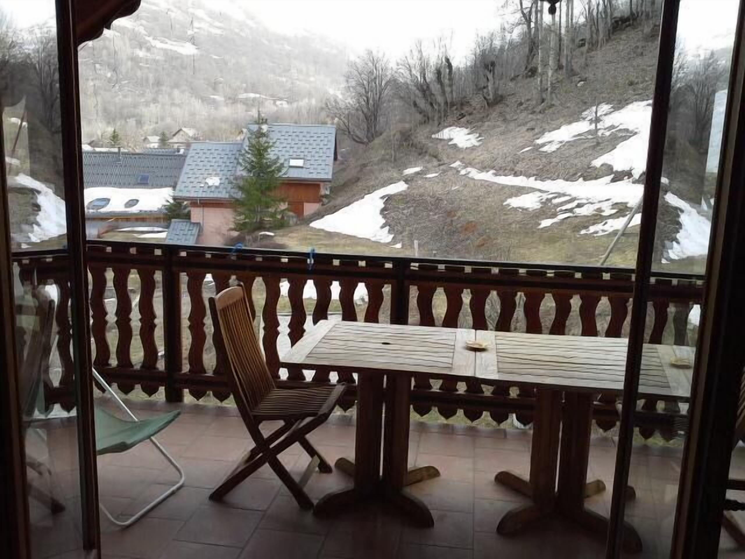 Un balcón con mesa y silla de madera, con vista a un paisaje nevado con casas y árboles.