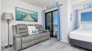 A room with a grey leather sofa, a white bed, a floor lamp, a painting of a pier, and a sign that reads 'BEACH'. - Royal Gulf Beach & Racquet 5302 - Beautiful Studio Gulf Front Unit! (Gulf Shores)