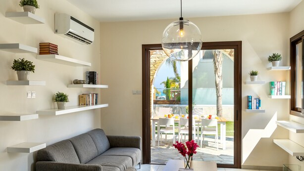 Una sala de estar moderna con un sofá gris, estanterías blancas con plantas, una lámpara colgante y vistas a un patio exterior.