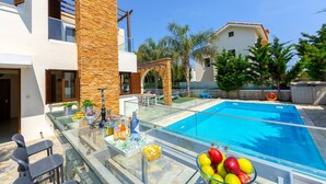 Un área de piscina exterior moderna con una mesa de vidrio, sillas y un frutero.