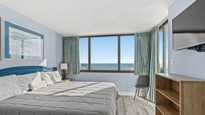 A bedroom with a large bed, a desk, a chair, a TV, a window with curtains, and a painting on the wall.