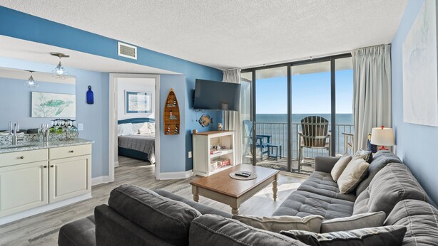 A living room with a sofa, coffee table, and a TV. There is a dining area with a table and chairs, and a kitchen with cabinets and a countertop.