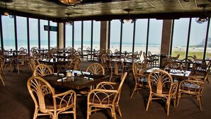 A restaurant with wicker chairs and tables, large windows, and a view of the beach.