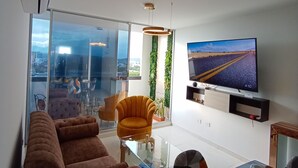Una sala de estar moderna con un sofá, una mesa de vidrio preparada para comer, un televisor montado en la pared y vistas de la ciudad a través de amplios ventanales.
