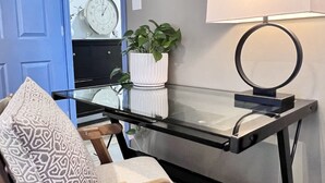 Una habitación de esquina con una puerta azul, un escritorio de vidrio con una lámpara, un reloj y una planta en maceta.