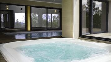 Piscina interior moderna com água cristalina, rodeada por grandes janelas que oferecem vista para árvores e um céu nublado.