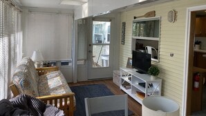 Een gezellige woonkamer met een bank, een televisie op een witte kast en een raam met zonwering.