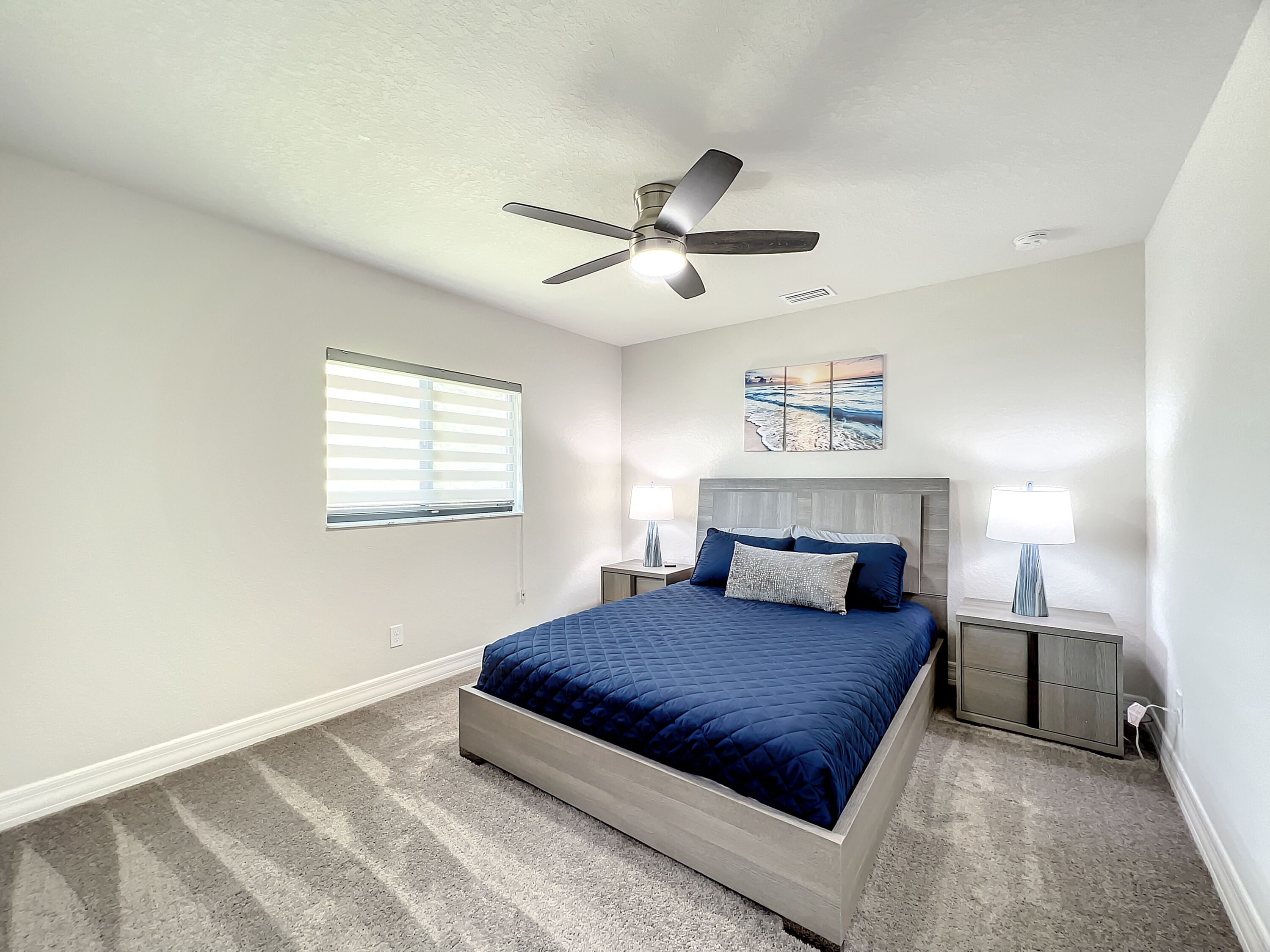 A bedroom with a bed, a ceiling fan, a window with blinds, a nightstand with a lamp, and a painting on the wall.