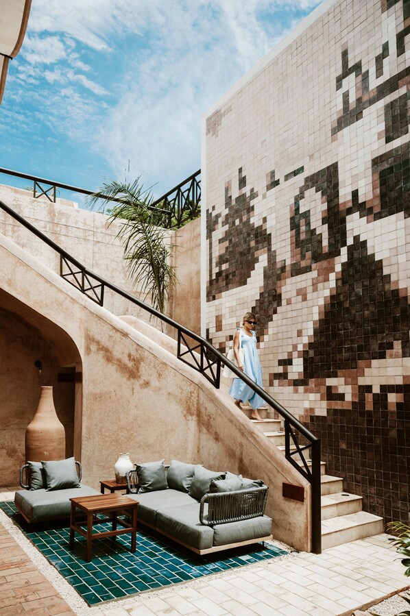 Un área exterior moderna con zona de estar, un sofá y una mesa, una escalera con barandilla y una pared de azulejos con diseño pixelado.