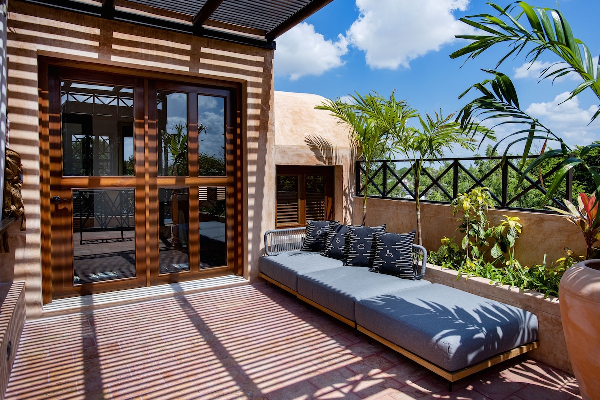 A modern outdoor seating area with a sofa and cushions, a wooden pergola, and a view of greenery and a clear sky.