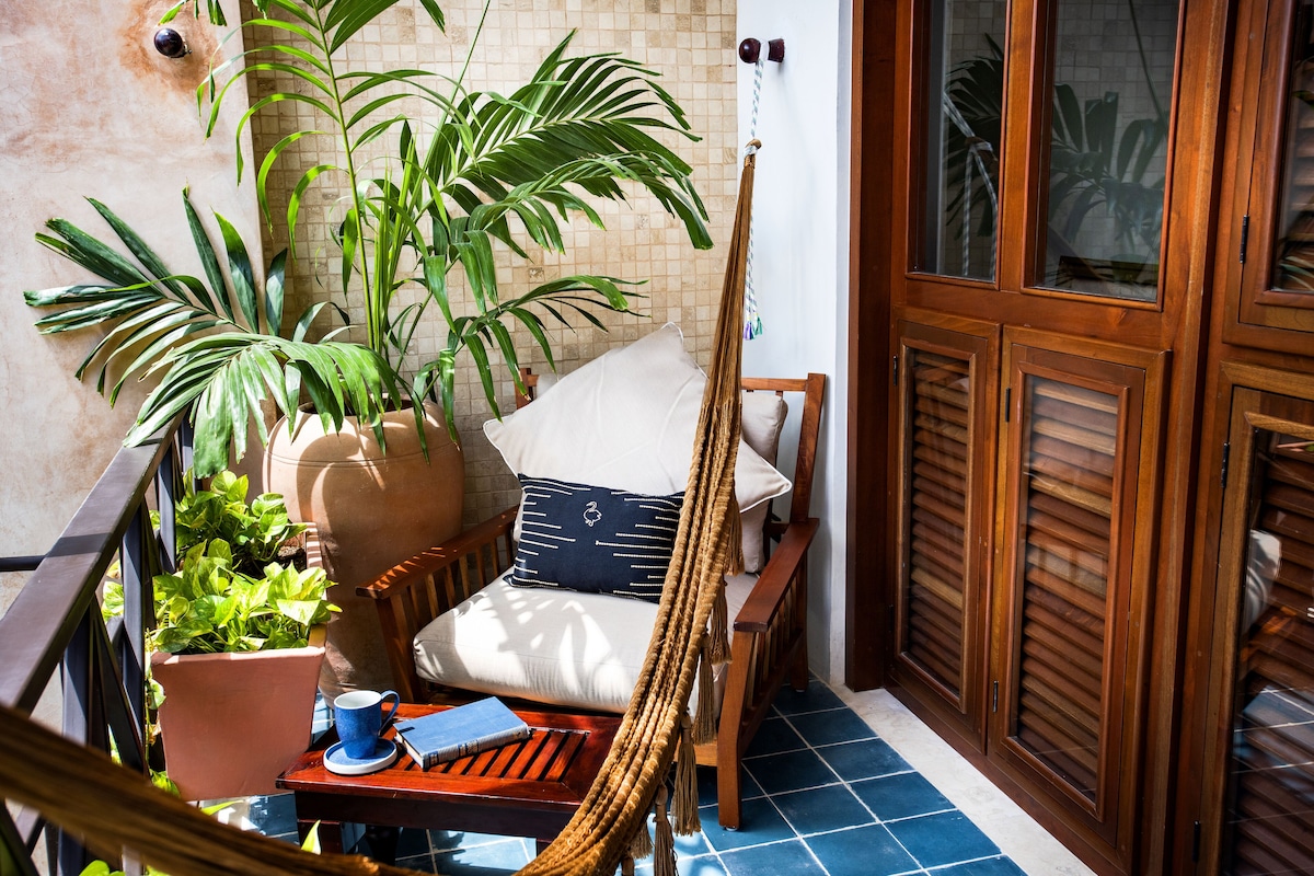 A balcony with a hammock, a potted plant, a chair with cushions, and a table with a cup and a notebook.