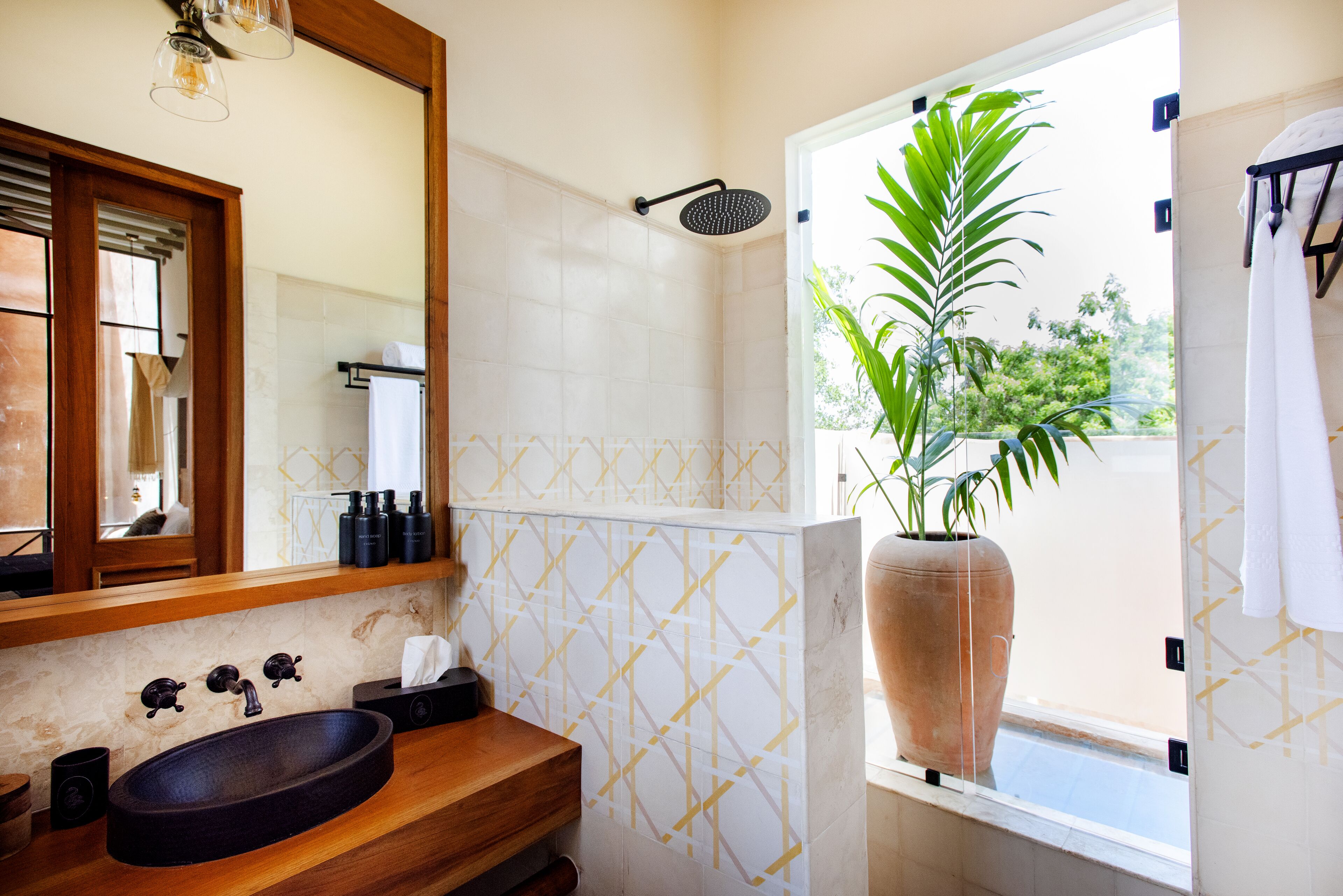 A bathroom with a large mirror, a wooden vanity, a black sink, a showerhead, and a potted plant by the window.