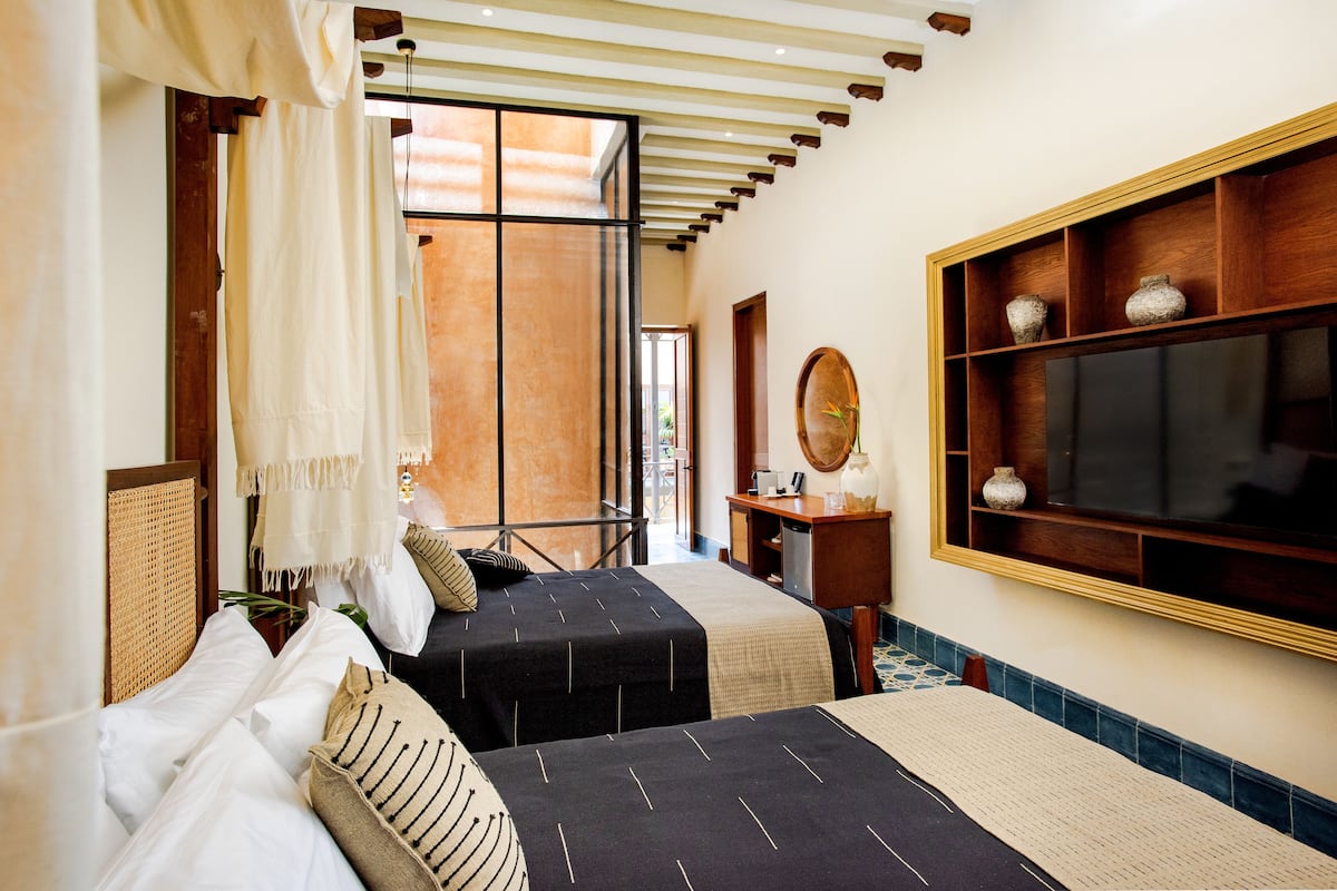 A hotel room with two beds, a wooden shelf with decorative items, a large mirror, and a view of the outside.