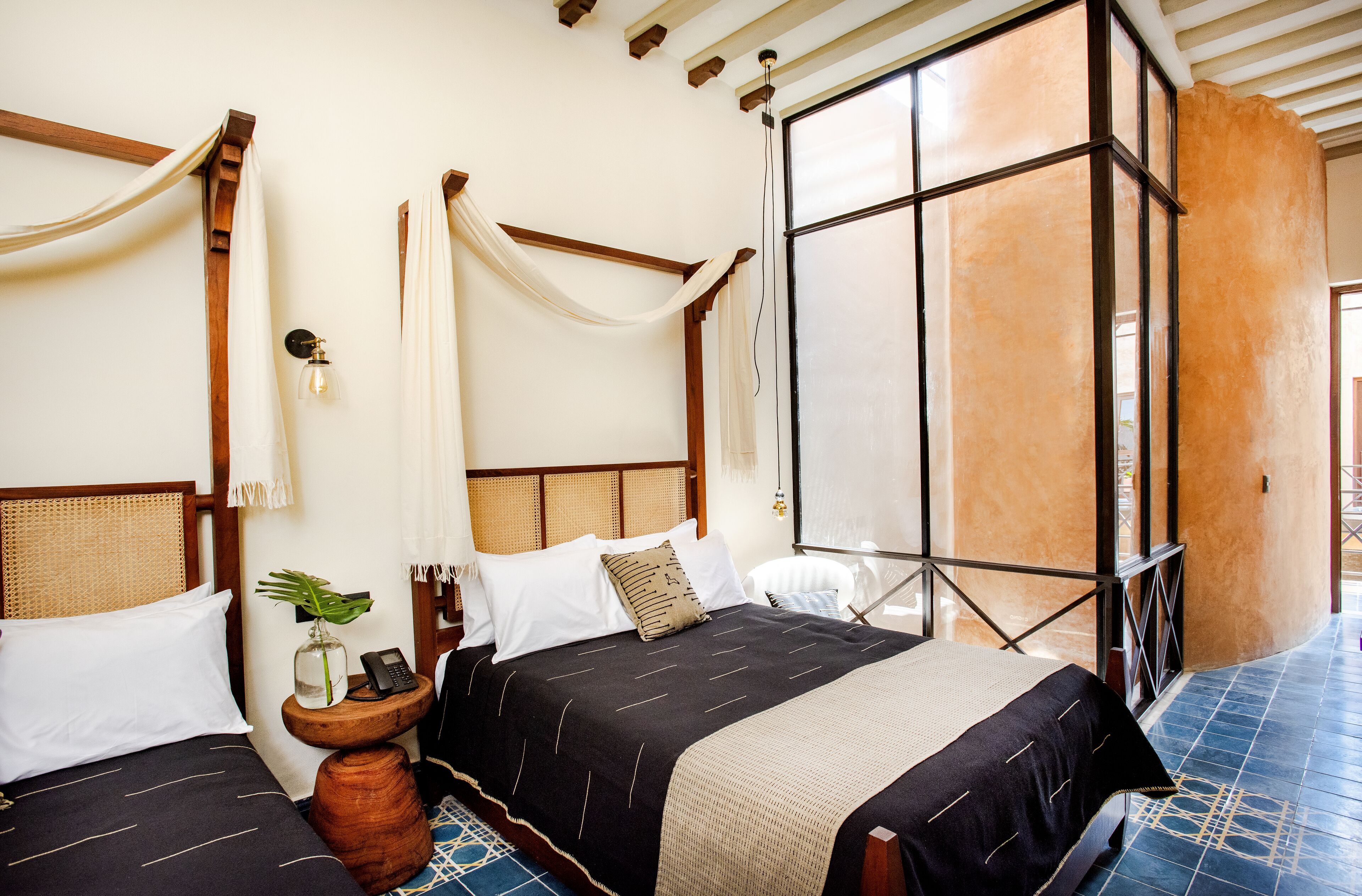 A room with two beds, each with a wooden headboard, a bedside table with a plant, and a large window with a view.