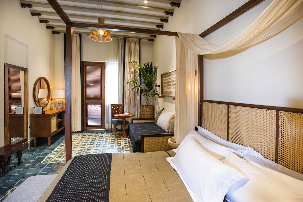 A hotel room with a large bed, a desk with a lamp, a chair, a small table, and a potted plant.