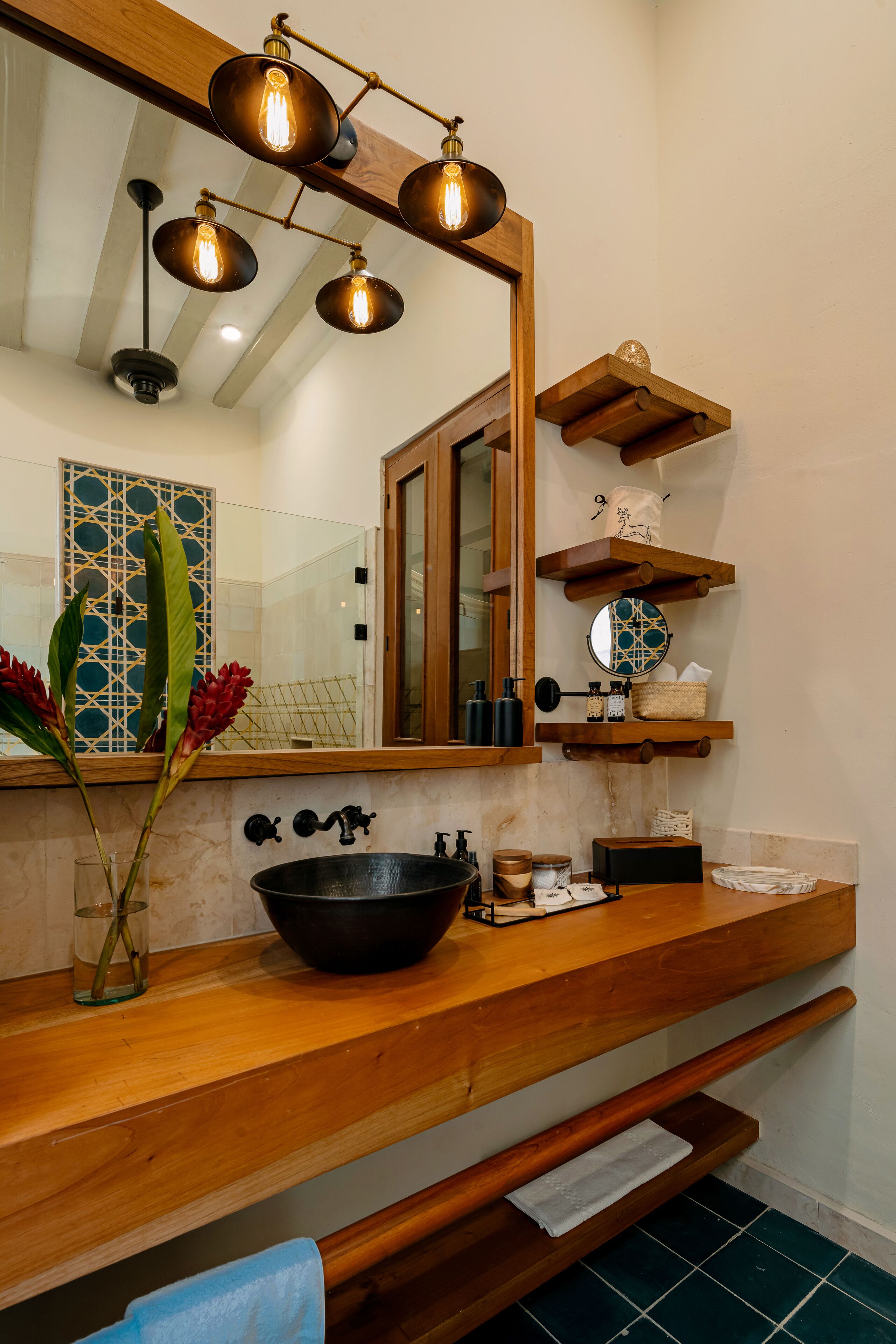 A bathroom with a wooden vanity, a large mirror, and a black basin. There are wooden shelves above the vanity and a towel rack below.