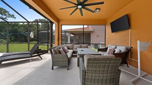 Une terrasse extérieure aménagée avec des meubles en osier, un ventilateur de plafond et un téléviseur fixé au mur.