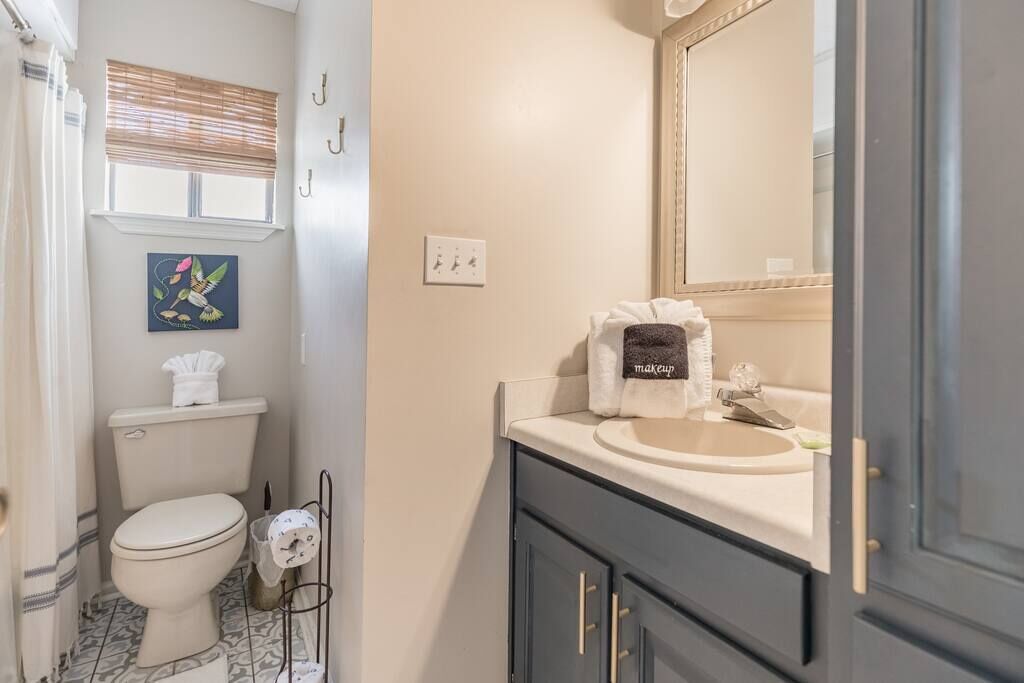 Ein Badezimmer mit Toilette, Waschbecken und Spiegel. Handtuchhalter mit Handtuch, ein Bild an der Wand und ein kleiner Mülleimer sind ebenfalls vorhanden.