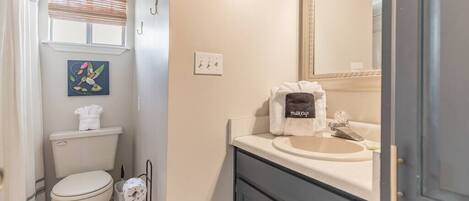 Ein Badezimmer mit Toilette, Waschbecken und Spiegel. Handtuchhalter mit Handtuch, ein Bild an der Wand und ein kleiner Mülleimer sind ebenfalls vorhanden.