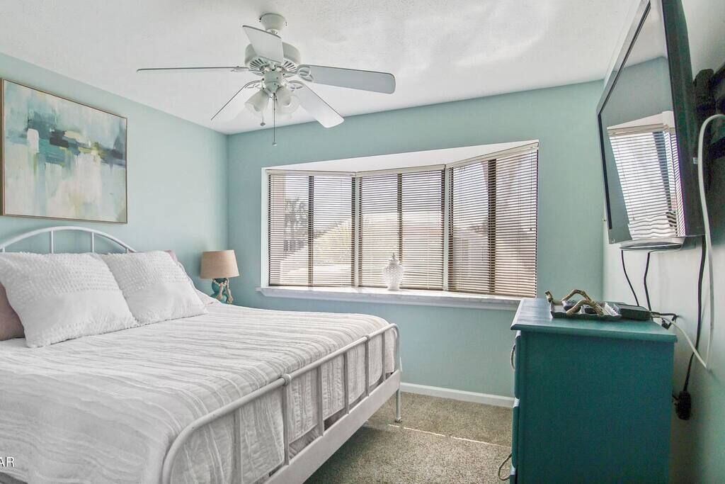 Un dormitorio con cama, ventilador de techo, ventana con persianas, cómoda y un cuadro en la pared.