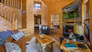 Ein Blockhaus-Interieur mit Kamin, einer Holztreppe und einer gemütlichen Sitzecke mit Sofa und Sesseln.