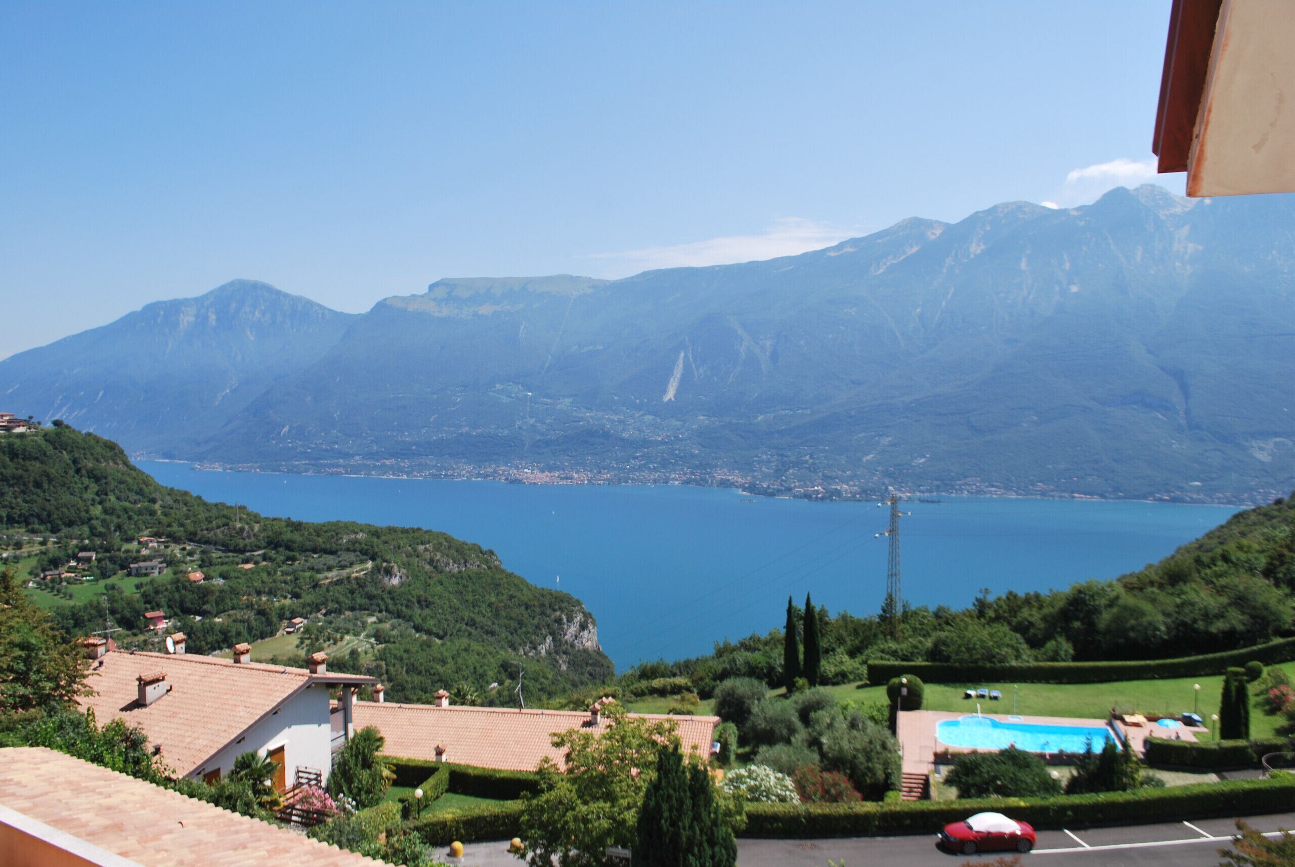 A view of a mountainous landscape with a lake, a swimming pool, and a red car parked in the foreground.