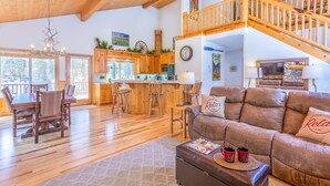 A spacious living area with a wooden ceiling, a dining table with chairs, a brown leather sofa with 'Relax' pillows, and a kitchen in the background. - ACER04 - Spacious Home, Hot Tub, Fireplace, Ping Pong, Foosball (Sunriver)