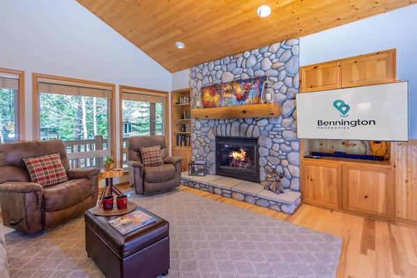 A cozy living room with a stone fireplace, wooden ceiling, and large windows overlooking a forest.