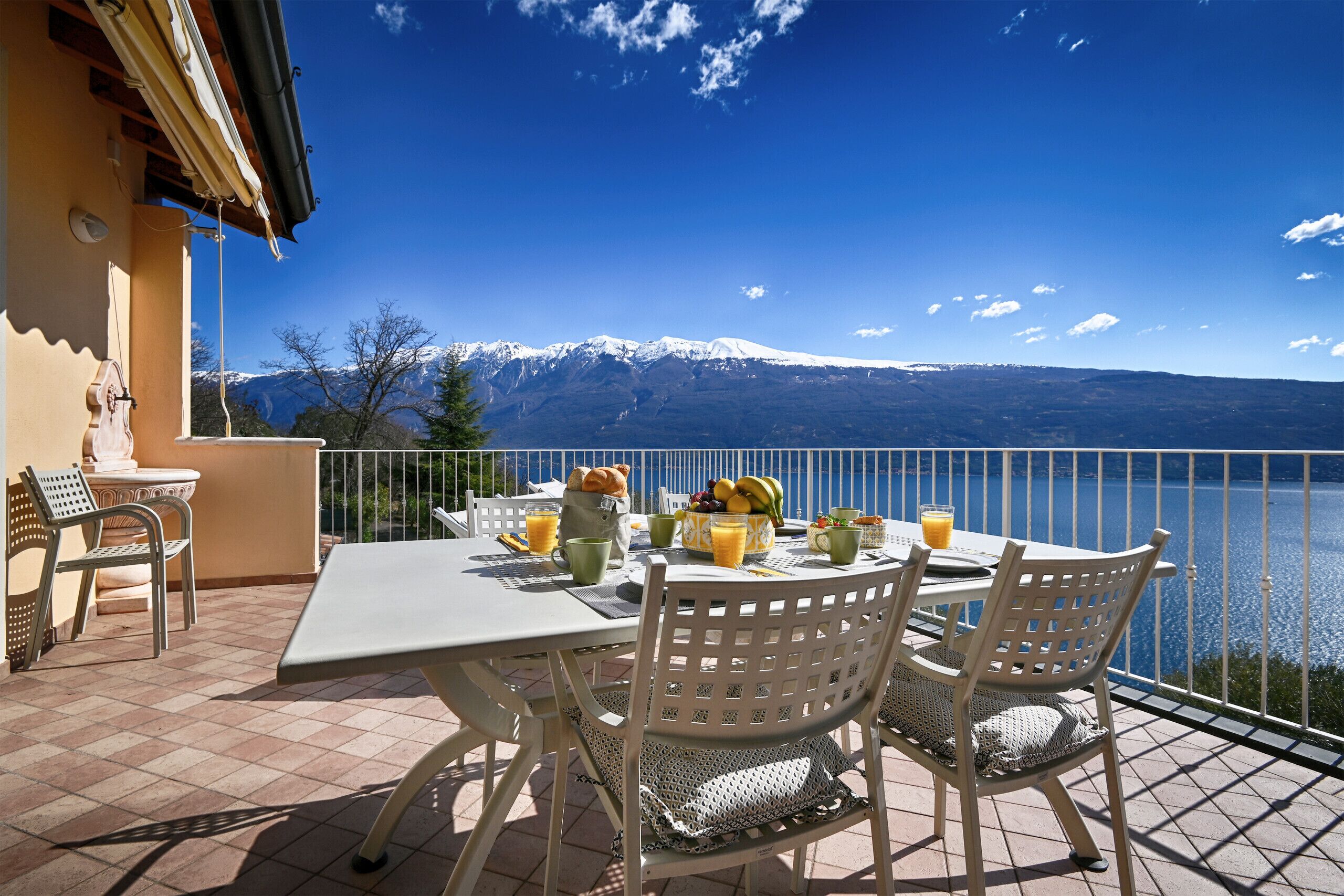 A terrace with a table set for breakfast, overlooking a lake and snow-capped mountains.