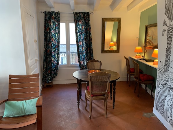 A dining area with a round table, four chairs, a window with curtains, a mirror, and two lamps on a shelf.